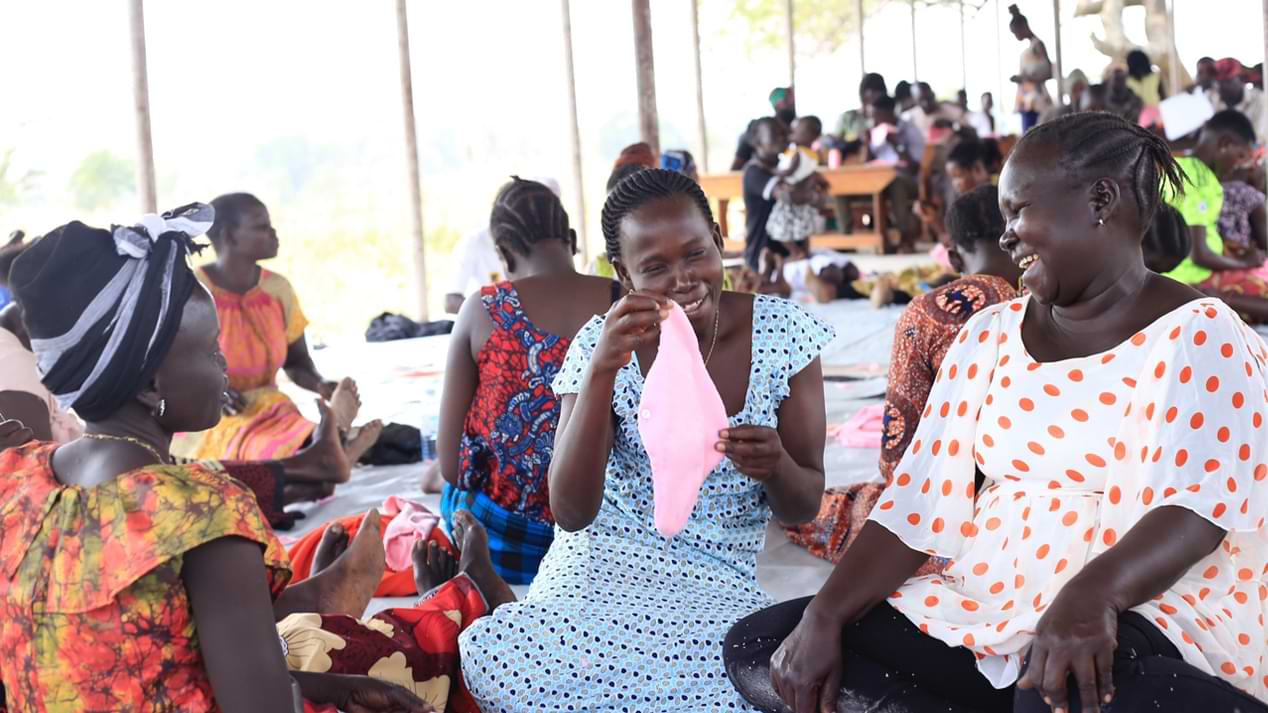 Eine Gruppe von Frauen in bunten Kleidern in einer Geflüchtetensiedlung in Uganda sieht sich eine Stoffbinde an und lacht.