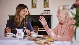 Junge Ehrenamtlerin des Besuchsdienstes sitzt mit einer Seniorin bei Kaffee und Kuchen zusammen.