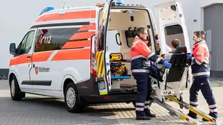 Malteser in Einsatzkleidung helfen einem gehbehindertem Mann in in ein Fahrzeug.