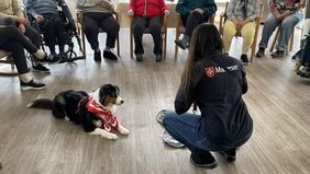 Umringt von Senioren auf Stühlen und Rollstühlen liegt ein Hund neben einer knienden Malteser Helferin.