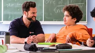 Malteser Schulbegleiter sitzt mit einem Jugendlichen im Rollstuhl im Klassenraum.