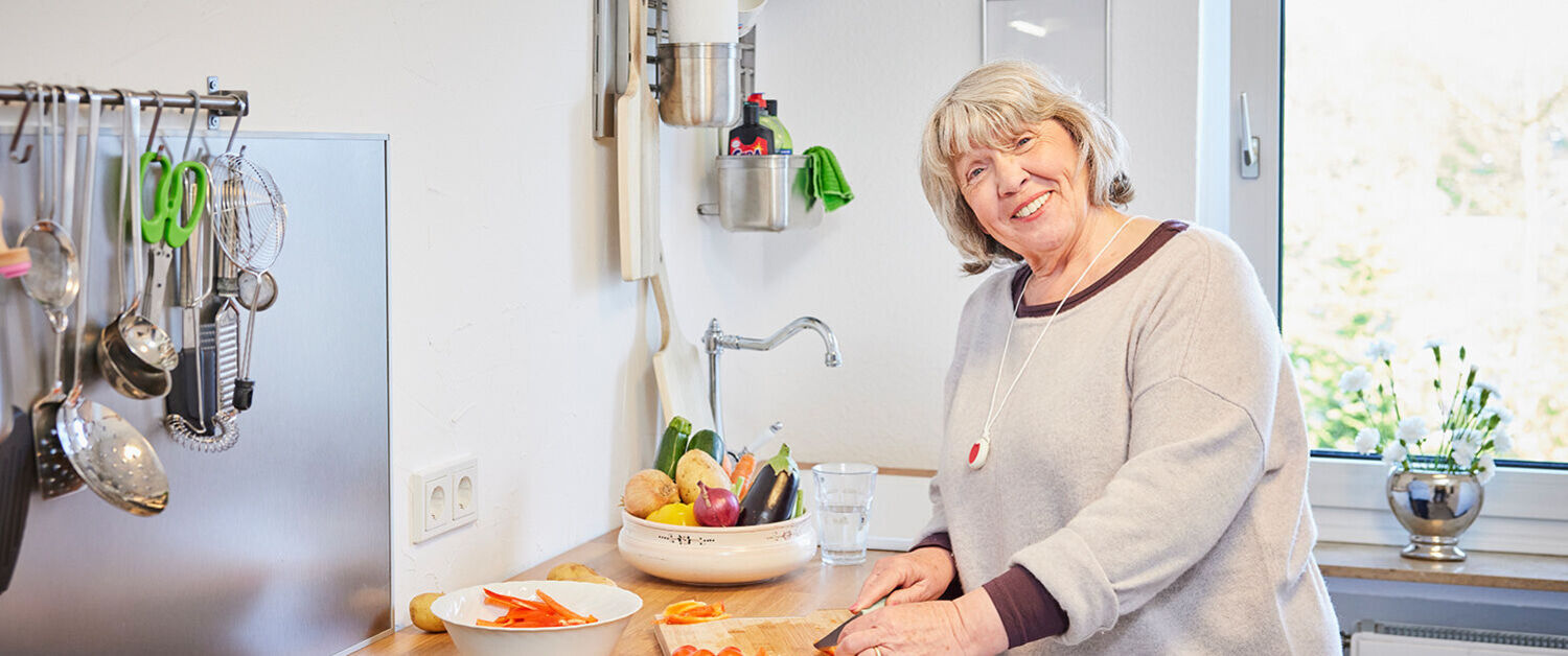 Eine Seniorin trägt den Hausnotrufknopf als Kette und kocht bei sich zuhause.