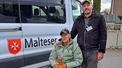 Ein Mann im Rollstuhl mit einem Blumentopf in der Hand, dahinter ein Mann in Malteserkleideung, dahinter ein Behindertentransportfahrzeug der Malteser.