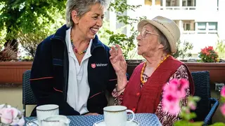 Eine Malteser Ehrenamtliche und eine ältere Dame unterhalten sich bei einer Tasse Kaffee.