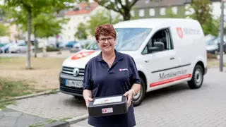 Mitarbeiterin des Malteser Menueservice trägt eine Warmhaltebox und lächelt in die Kamera.