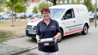 Mitarbeiterin des Malteser Menueservice trägt eine Warmhaltebox und lächelt in die Kamera.