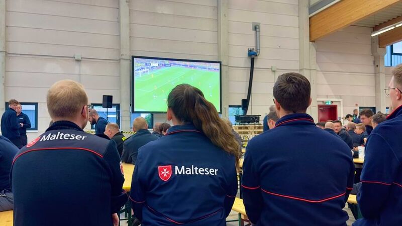 Menschen in Einsatzkleidung des Malteser Hilfsdienstes schauen ein Fußballspiel auf einem Fernseher