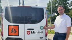 Ein Mann in Malteserkleidung steht bei einem Malteser Kleinbus und zeigt auf ein Dekra-Siegel.