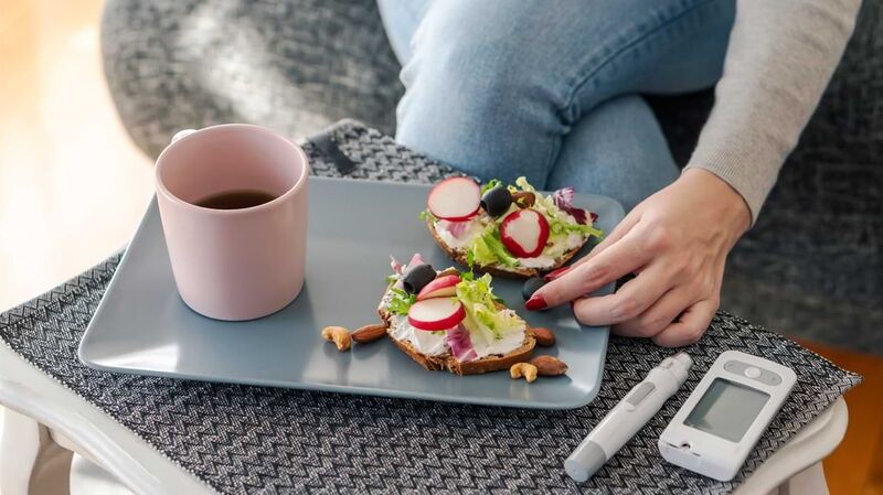 Eine Frau greift nach einem belegten Brot auf einem Tablet; daneben liegen Geräte zum Messen des Blutzuckerspiegels.