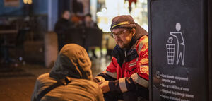 Ein Malteser Mitarbeiter spricht zu einem obdachlosen Mann, der neben einem Mülleimer an der Straße sitzt