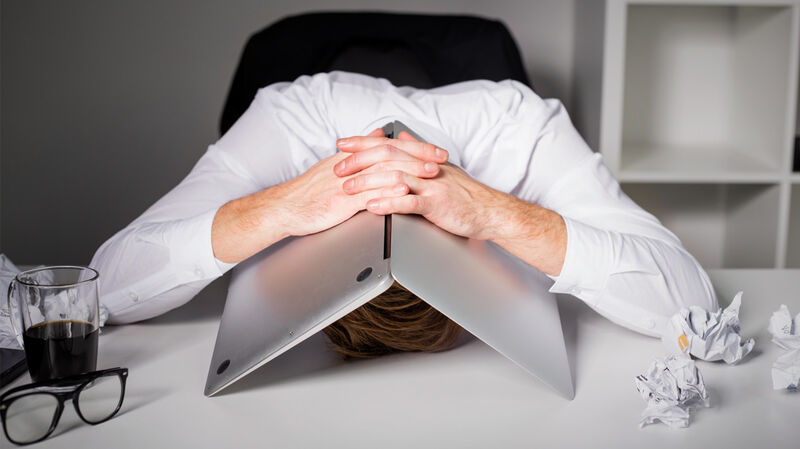 Ein Mann am Schreibtisch, versteckt seinen Kopf unter einem Laptop 