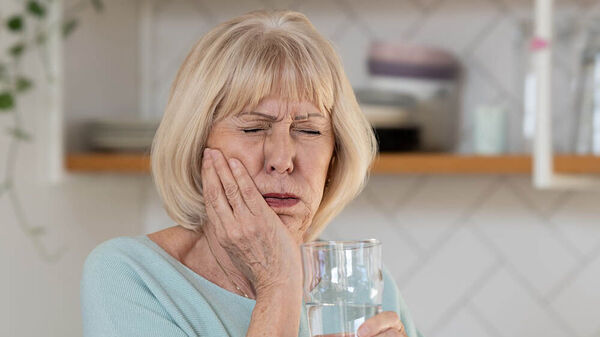 Eine ältere Frau, mit einem Glas in der Hand, verzieht schmerzerfüllt das Gesicht und hält sich die Wange.